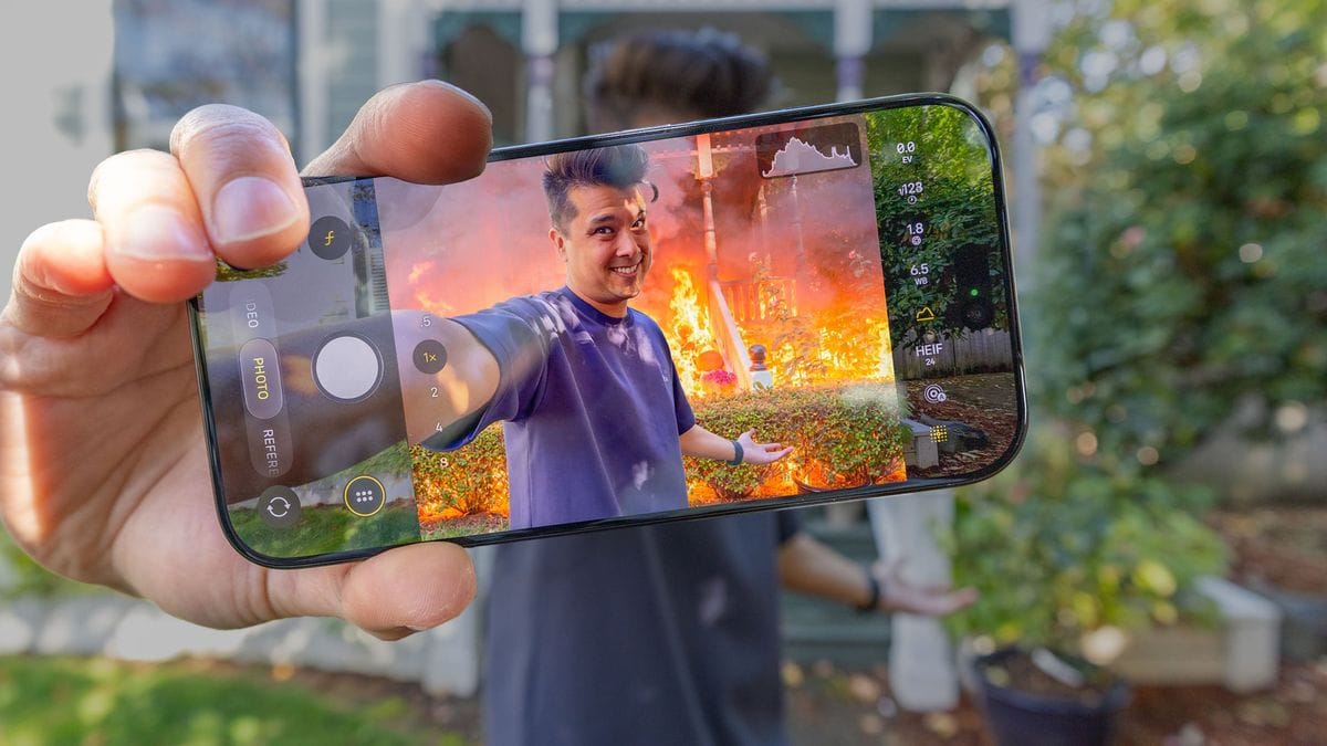 A hand holds a smartphone displaying a camera interface, showing a man smiling in front of a house engulfed in flames.