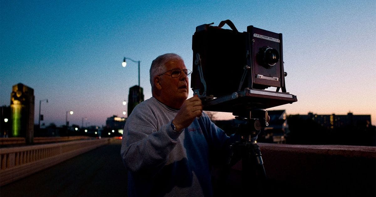 An older man with glasses stands on a bridge at dusk, adjusting a large-format bellows camera mounted on a tripod.